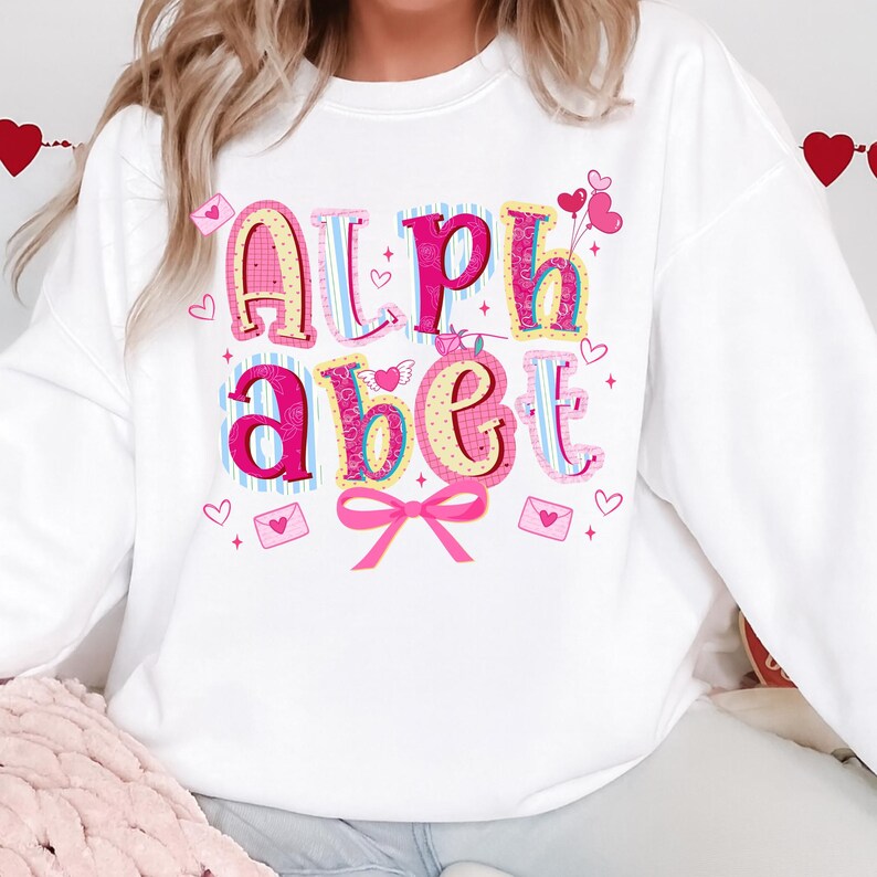 May include: White sweatshirt with the word "Alphabet" in colorful, decorative letters. The letters are pink, yellow, and blue, with hearts and a pink bow. The sweatshirt has a Valentine's Day theme.