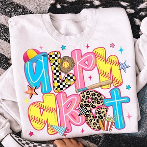 May include: White sweatshirt with a colorful softball-themed graphic. The design features the words "Girl's Market" in large, playful letters with baseball and softball elements, including a glove, bat, and popcorn.