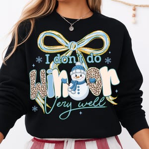 May include: Black sweatshirt with a winter-themed graphic. The design features a snowman wearing a blue hat and scarf, with the text "I don't do winter very well." The word "winter" has a leopard print pattern.