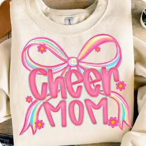 May include: Cream-colored sweatshirt with a large pink bow and the words "Cheer MOM" in pink, outlined in a darker pink. The bow has rainbow stripes and small pink flowers are scattered around the design.