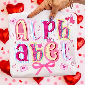 May include: White t-shirt with the word "ALPHABET" in a colorful, whimsical design. The letters are pink, blue, and yellow, with heart and envelope accents. A pink bow is at the bottom. The background includes red heart-shaped balloons.