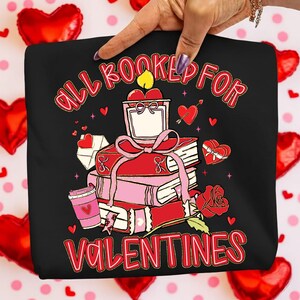 May include: Black graphic tee with the text "ALL BOOKED FOR VALENTINES" in red script. The design features a stack of books tied with a pink ribbon, a candle, a rose, and hearts. A coffee cup and a heart-shaped gift box are also included.