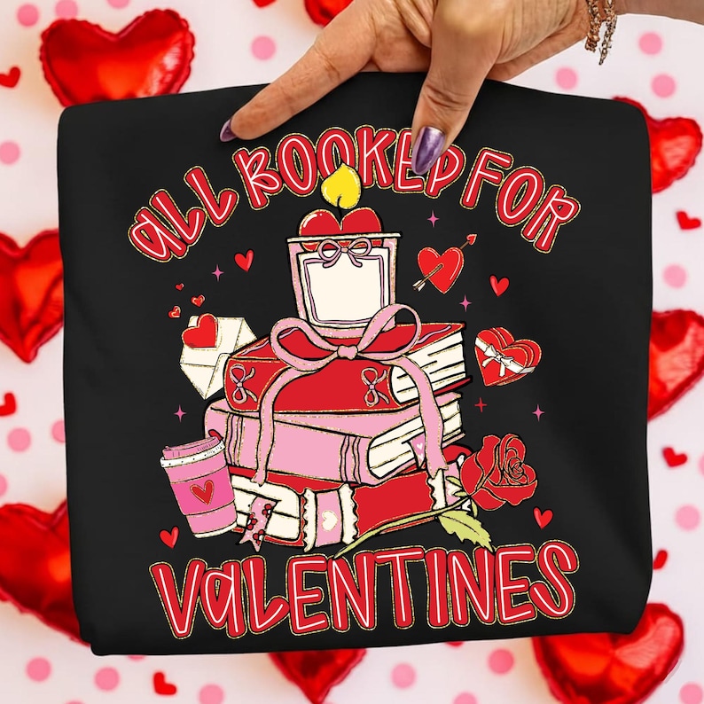 May include: Black graphic tee with the text "ALL BOOKED FOR VALENTINES" in red script. The design features a stack of books tied with a pink ribbon, a candle, a rose, and hearts. A coffee cup and a heart-shaped gift box are also included.