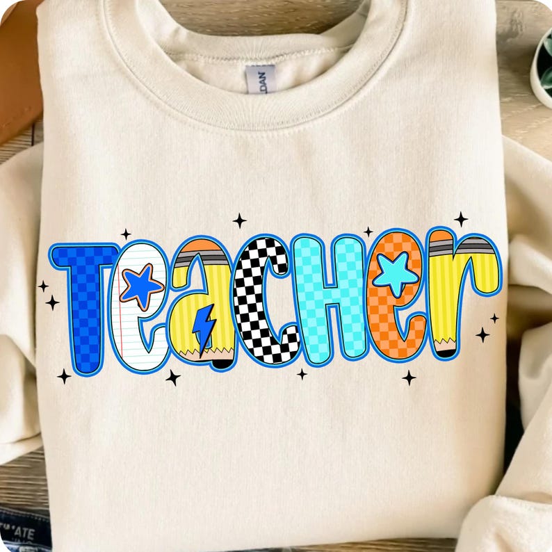 May include: Cream-colored sweatshirt with the word "Teacher" in a colorful, playful design. The letters are outlined in blue, with various patterns and illustrations, including a star, pencil, and lightning bolt.