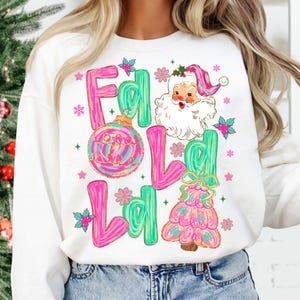 May include: White sweatshirt with a festive design. The design includes the words "Fa La La" in pink and green, along with a Santa Claus illustration, Christmas ornaments, and a Christmas tree.