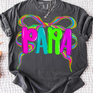 May include: Dark gray t-shirt with the word "PAPA" in bright pink, teal, and green letters. A colorful rainbow bow design surrounds the word. The shirt is made of a soft material.