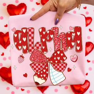 May include: A light pink sweatshirt with the word "Alpha" in red, decorated with hearts, a bow, and a pencil. The bow has a checkered pattern and a leopard print. A strawberry and the letters "XOXO" are also featured.