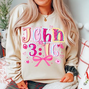 May include: A cream-coloured sweatshirt featuring the text "John 3:16" in a colourful, decorative font. The design incorporates hearts, a bow, and other Valentine's Day themed details. The sweatshirt is a relaxed, comfortable style.