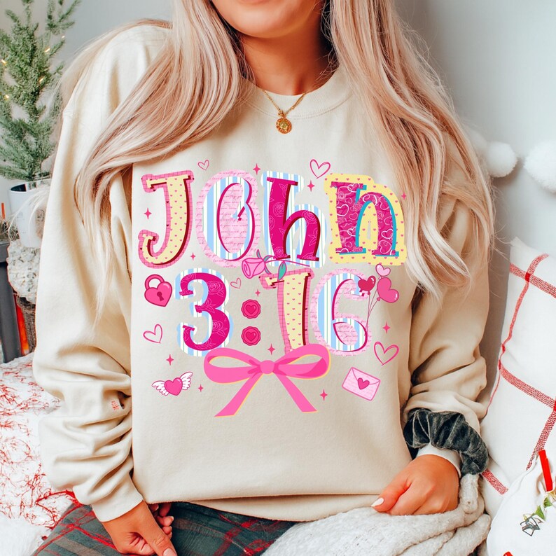 May include: A cream-coloured sweatshirt featuring the text "John 3:16" in a colourful, decorative font. The design incorporates hearts, a bow, and other Valentine's Day themed details. The sweatshirt is a relaxed, comfortable style.