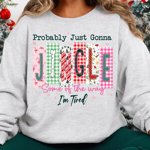 May include: A light gray sweatshirt with the text "Probably Just Gonna Jingle Some of the way I'm Tired" in a colorful, festive design. The word "JINGLE" is in a block letter style with various patterns and colors, perfect for the holiday season.
