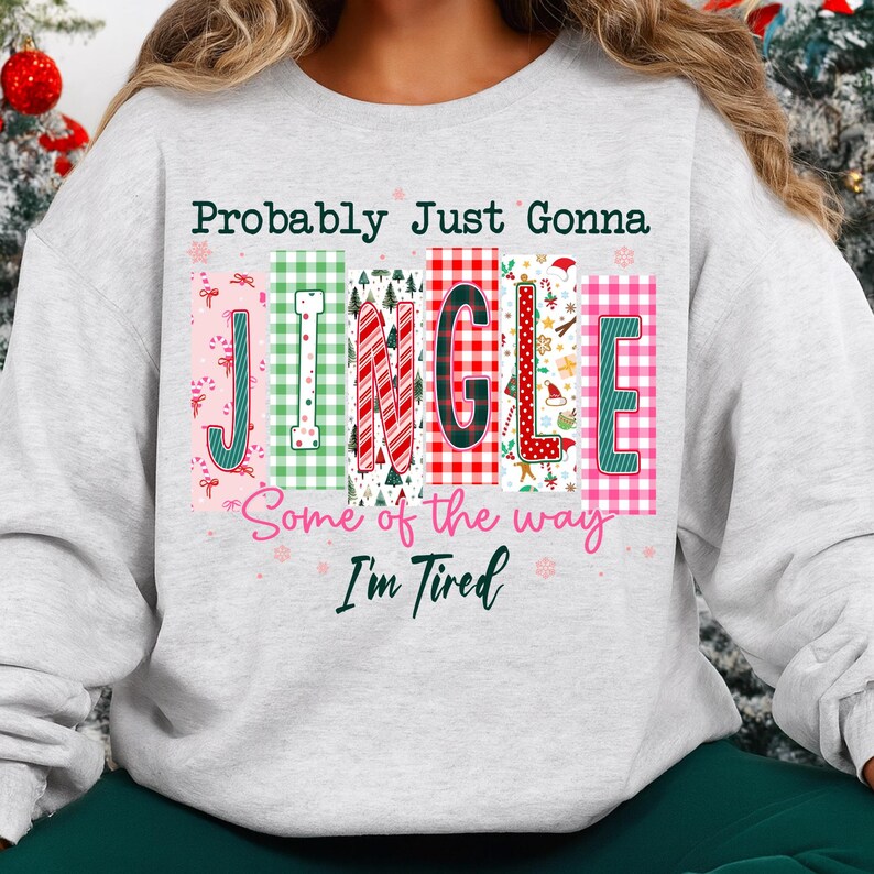 May include: A light gray sweatshirt with the text "Probably Just Gonna Jingle Some of the way I'm Tired" in a colorful, festive design. The word "JINGLE" is in a block letter style with various patterns and colors, perfect for the holiday season.