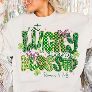 May include: Cream-colored sweatshirt with the text "not LUCKY simply BLESSED" in green, pink, and gold lettering. The design includes shamrocks and pink bows. The Bible verse Roman 4:7-8 is also printed on the shirt.