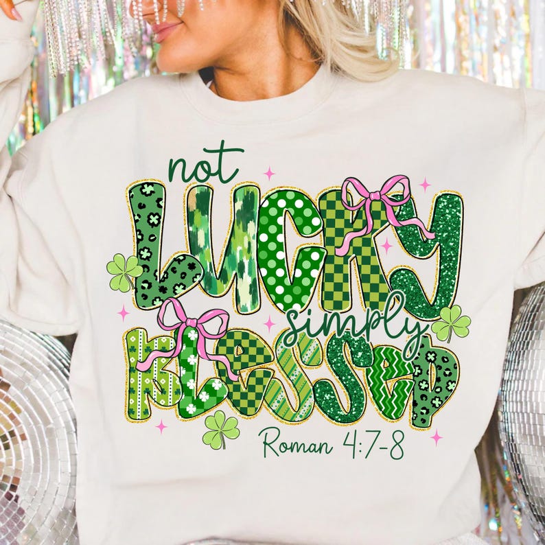 May include: Cream-colored sweatshirt with the text "not LUCKY simply BLESSED" in green, pink, and gold lettering. The design includes shamrocks and pink bows. The Bible verse Roman 4:7-8 is also printed on the shirt.