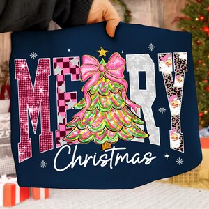 May include: Navy blue Christmas-themed graphic tee with the words "MERRY Christmas" in a festive design. The "M" is pink with sequins, the "E" is checkered, and the "R" and "Y" are white. A colorful Christmas tree with a pink bow is in the center.