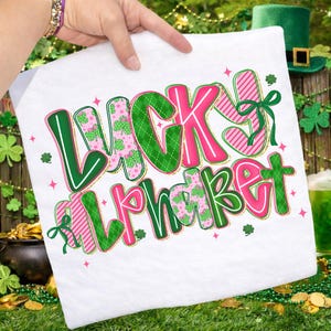 Pu&ograve; includere: Tessuto bianco con la scritta "LUCKY ALPHABET" in grandi lettere colorate. Le lettere sono verdi e rosa, con motivi a trifoglio e a righe. Fiocchi verdi e dettagli dorati completano il design festivo, perfetto per San Patrizio.