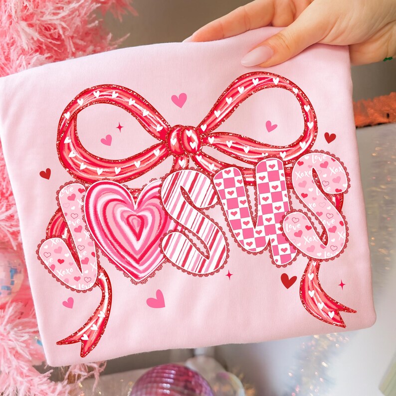 May include: A light pink t-shirt featuring the word "JESUS" in a decorative font with heart and bow accents. The letters are filled with patterns of hearts and stripes. The design includes a large bow and several small hearts.