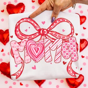 May include: White t-shirt with a large red bow and the word "MIMI" in pink and red lettering. The "O" in MIMI is a heart shape. Small hearts and decorative ribbons are also featured.