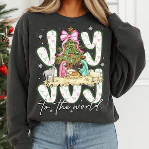 May include: Dark gray sweatshirt with a Christmas-themed graphic. The design features the word "JOY" in white letters with a green and pink bow pattern, a Christmas tree, and a nativity scene. The phrase "to the world" is below the word "JOY".