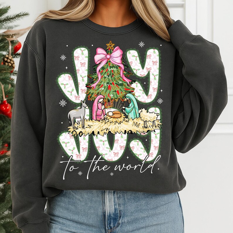 May include: Dark gray sweatshirt with a Christmas-themed graphic. The design features the word "JOY" in white letters with a green and pink bow pattern, a Christmas tree, and a nativity scene. The phrase "to the world" is below the word "JOY".