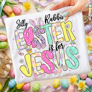 May include: White t-shirt with the words "Silly Rabbit Easter is for Jesus" in colorful, playful lettering. The design includes Easter eggs, a bunny, and a rainbow, perfect for Easter celebrations.