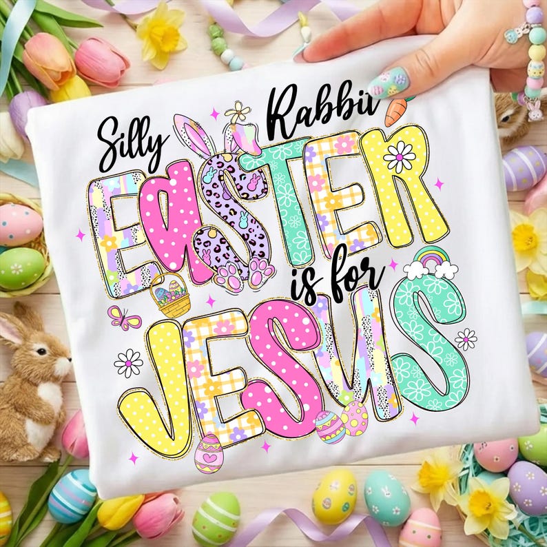 May include: White t-shirt with the words "Silly Rabbit Easter is for Jesus" in colorful, playful lettering. The design includes Easter eggs, a bunny, and a rainbow, perfect for Easter celebrations.
