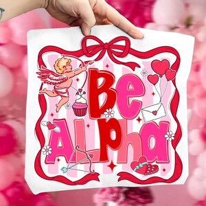 May include: A white tote bag with a Valentine's Day design. The design features the words "Be Alpha" in red and pink, along with illustrations of Cupid, hearts, flowers, and a bow and arrow. A red ribbon frames the design.