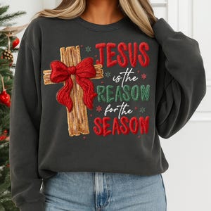 May include: Dark gray sweatshirt featuring a wooden cross with a red bow and the text "JESUS is the REASON for the SEASON" in red and green. The design includes snowflakes, perfect for the Christmas holiday.
