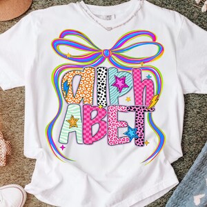 May include: White t-shirt with a colorful "ALPHABET" graphic. The letters feature various patterns, including leopard print, stripes, and checkered designs. A large bow and stars surround the text, creating a vibrant and playful design.