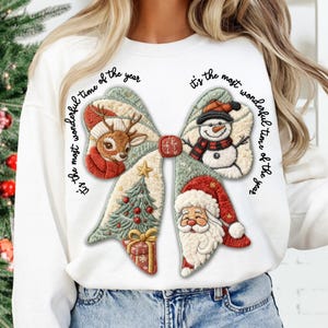 May include: White long-sleeve sweatshirt featuring a large bow design with Christmas-themed embroidered elements. The bow includes a reindeer, snowman, Christmas tree, Santa Claus, and the text "It's the most wonderful time of the year."