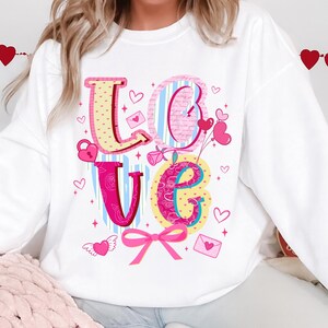 以下が含まれることがあります： カラフルで気まぐれなデザインの「LOVE」という文字がプリントされた白いスウェットシャツ。文字はピンク、黄色、青で、ハート、リボン、その他のバレンタインデーをテーマにしたグラフィックで飾られています。キュートでフェスティバルなトップス。