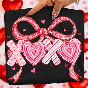 May include: Black sweatshirt featuring a red bow and the text "XOX" in a heart design. The bow and hearts have a pink and white colour scheme, with small hearts scattered around the design. A hand is holding the sweatshirt.