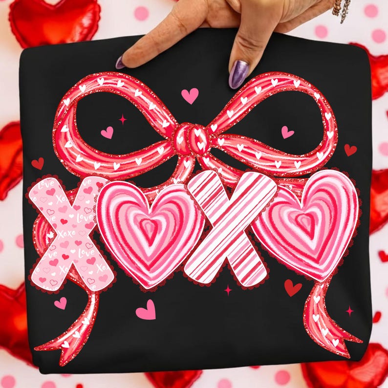 May include: Black sweatshirt featuring a red bow and the text "XOX" in a heart design. The bow and hearts have a pink and white colour scheme, with small hearts scattered around the design. A hand is holding the sweatshirt.
