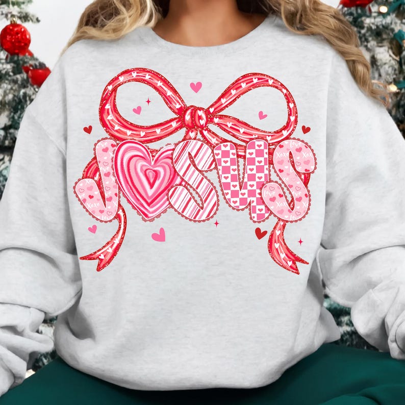 May include: A light gray sweatshirt featuring a large, decorative design with the word "JESUS" in pink and white. The design includes a bow, hearts, and ribbons, creating a festive and loving theme. The sweatshirt is a comfortable, casual style.