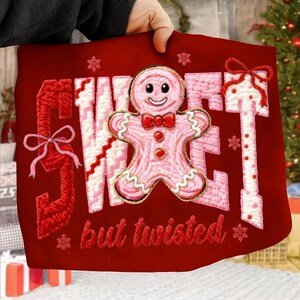 May include: Red Christmas-themed pillow with the words "SWEET but twisted" embroidered in red and pink yarn. A pink gingerbread man with a red bow tie is in the center. Snowflakes and bows add to the festive design.