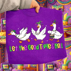 May include: A purple sweatshirt featuring three white geese wearing Mardi Gras hats and necklaces. The geese are dancing, and the text "Let the Good Times Roll" is displayed in green, yellow, and purple.