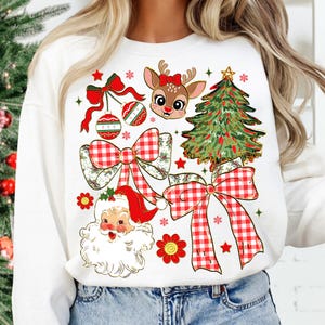 May include: White sweatshirt with a Christmas-themed design. The design features a reindeer with a red bow, a Christmas tree, Santa Claus, red and white plaid bows, baubles, and snowflakes. The graphic is colourful and detailed.