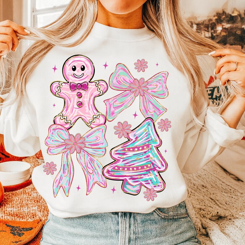 May include: White sweatshirt with a pink gingerbread man, bows, and a Christmas tree design. The gingerbread man has a bow tie and buttons. The bows and tree have pink, blue, and gold accents. Pink flowers and stars complete the festive design.