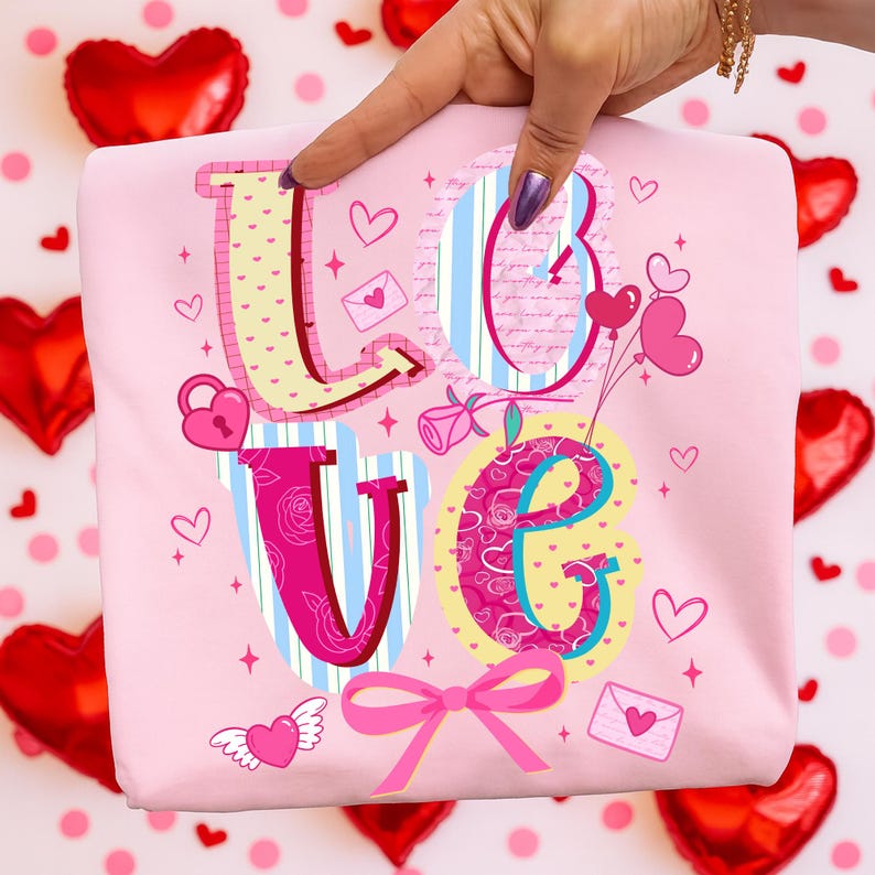 以下が含まれることがあります： カラフルな装飾フォントで「LOVE」という文字が描かれた、薄いピンク色のスウェットシャツ。文字はハート、バラ、その他のバレンタインデーをテーマにしたグラフィックで飾られています。下部にはピンクのリボンがあります。