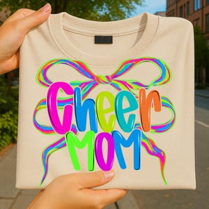 May include: A beige sweatshirt with the words "Cheer Mom" in colorful, bubble letters. A large, rainbow-colored bow design is above the text. The sweatshirt is held up by hands.