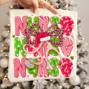 May include: A cream-colored t-shirt featuring the word "NURSE" in a festive design. The letters are in pink and green, with a bow and Santa hat. A pink stethoscope is also featured in the design.