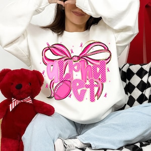 May include: White sweatshirt with a pink bow and the words "Fight Get" in pink checkered letters. A pink football is also featured in the design. Pink ribbons are scattered around the design.