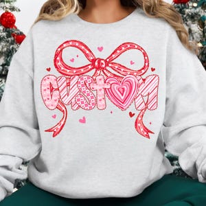 May include: A light gray sweatshirt with a large pink bow and the word "CUSTOM" in a heart-shaped design. The letters are decorated with pink hearts and stripes. Small pink hearts are scattered around the design, creating a festive look.