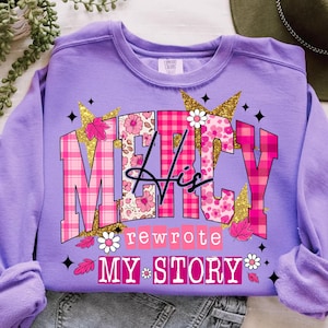 Op de afbeelding: Een lila sweatshirt met de tekst "MERCY His rewrote MY STORY" in een roze bloemen- en ruitpatroon. Het ontwerp bevat gouden glitteraccenten, bloemen en bladeren. Het sweatshirt is opgevouwen.