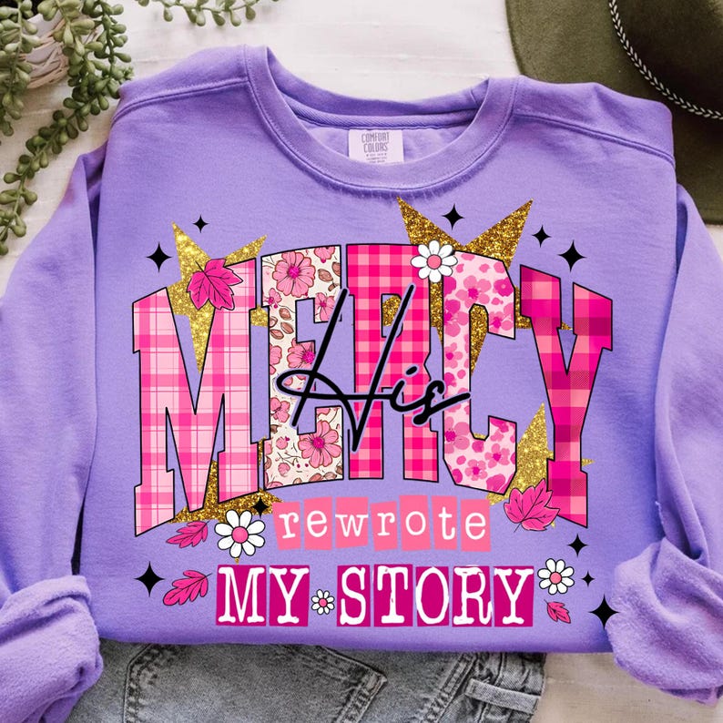 Op de afbeelding: Een lila sweatshirt met de tekst "MERCY His rewrote MY STORY" in een roze bloemen- en ruitpatroon. Het ontwerp bevat gouden glitteraccenten, bloemen en bladeren. Het sweatshirt is opgevouwen.