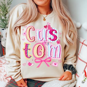 May include: A beige sweatshirt with the word "Custom" in a colorful, decorative font. The letters are pink, blue, and yellow, with heart and bow accents. The sweatshirt is a casual style.