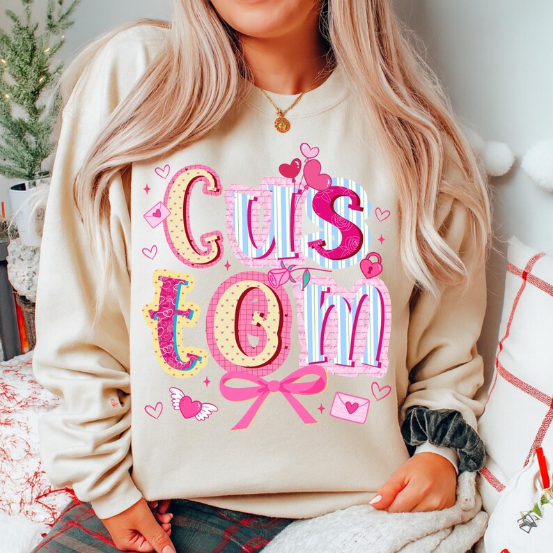 May include: A beige sweatshirt with the word "Custom" in a colorful, decorative font. The letters are pink, blue, and yellow, with heart and bow accents. The sweatshirt is a casual style.