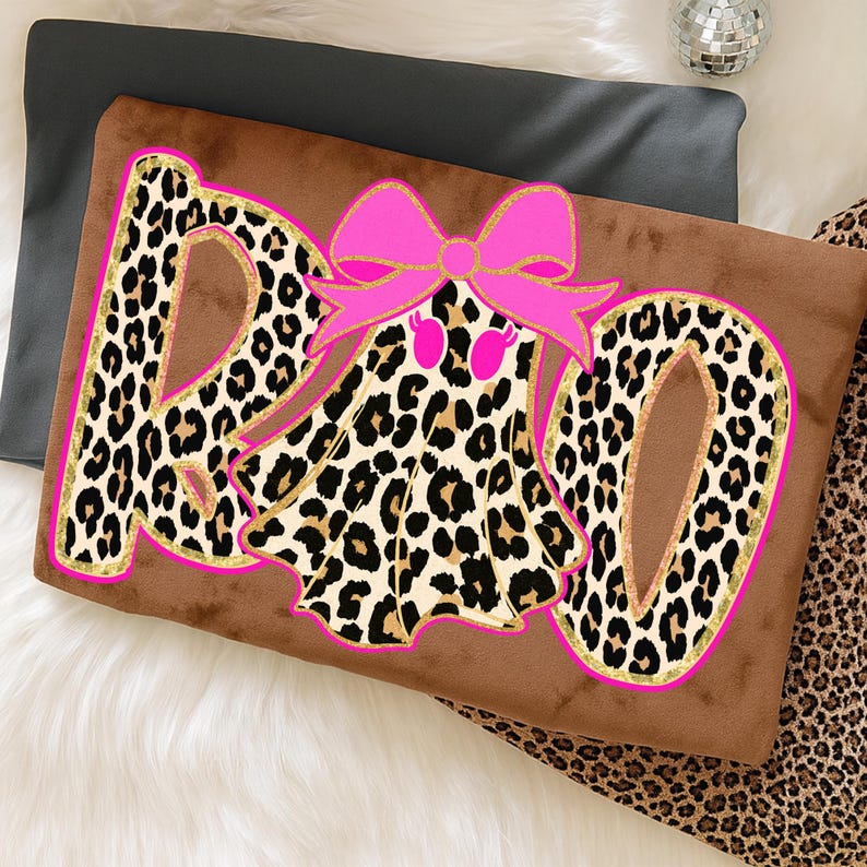 May include: Brown decorative pillow with the word "Boo" in leopard print letters outlined in pink. A pink bow with gold glitter accents is above a ghost-shaped design with a leopard print pattern.