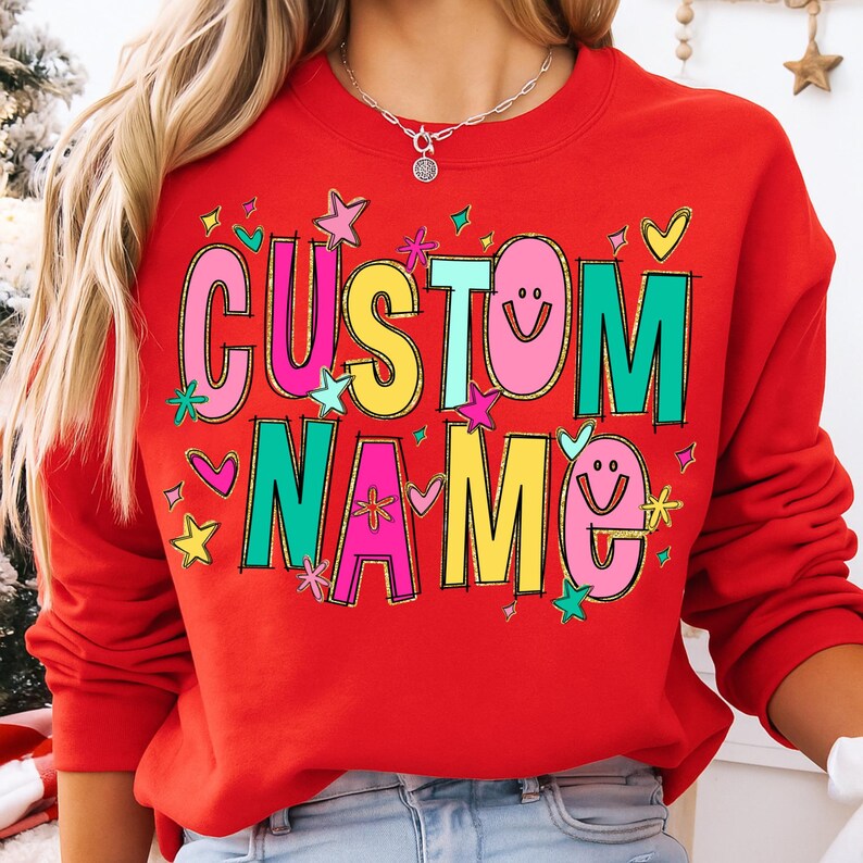 May include: A red sweatshirt with the words "CUSTOM NAME" in colorful, playful lettering. The design includes stars, hearts, and smiley faces. The sweatshirt is a vibrant, eye-catching piece.