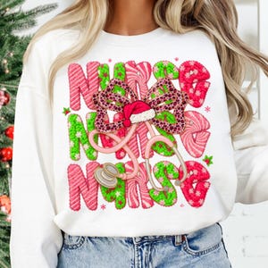 May include: White sweatshirt with the word "NURSE" in pink and green candy cane-style letters. A pink stethoscope and a bow with a Santa hat are in the center. The bow has a leopard print design.