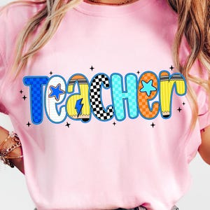 May include: Pink t-shirt with the word "Teacher" in a colorful, playful font. The letters are outlined in blue, orange, and yellow, with star and pencil details. The shirt is a light pink color.